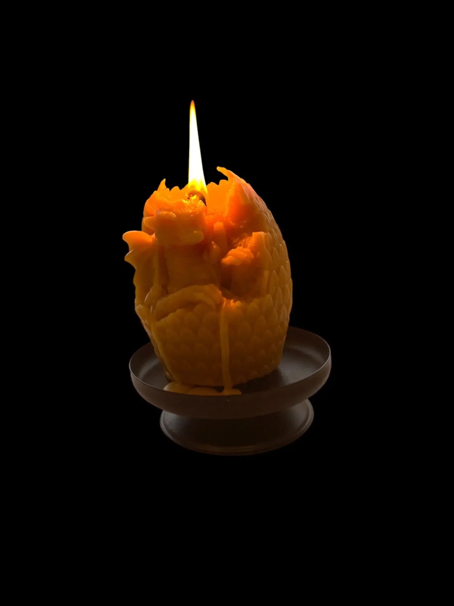 Lit orange dragon egg candle with detailed scales melting on a dark stand against black background