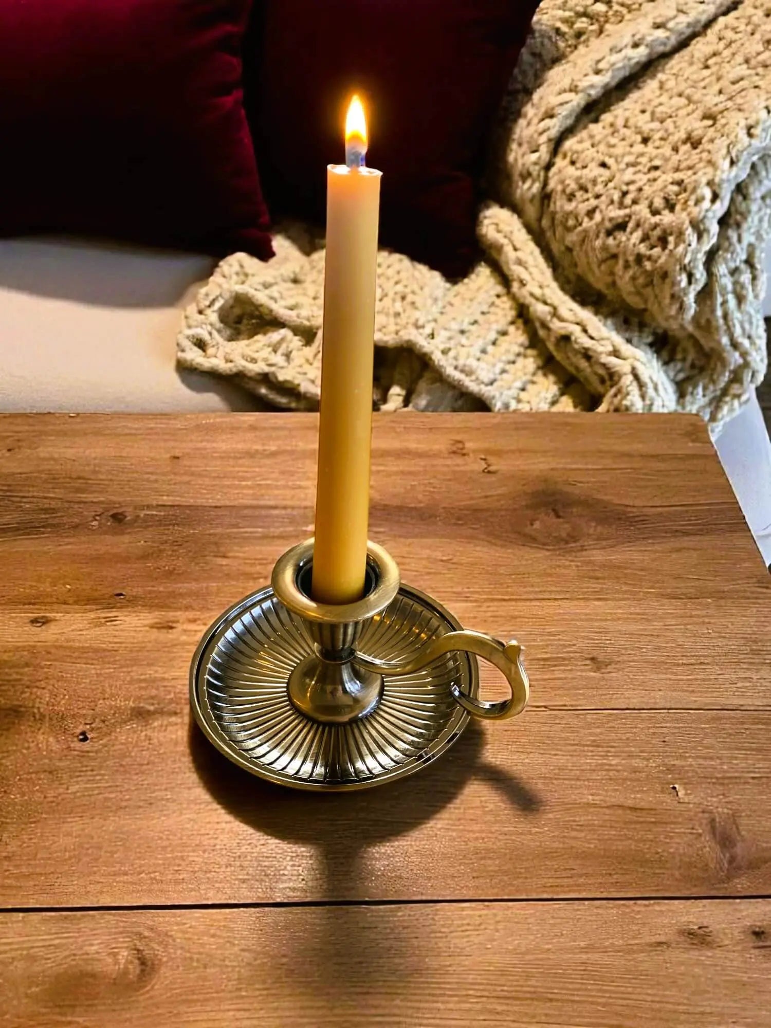 Lit handmade beeswax taper candle in vintage brass holder on wooden table with cozy knit blanket and pillow background