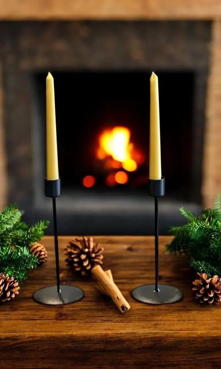 Pair of handmade beeswax taper candles on black holders by rustic wooden table with pinecones and greenery