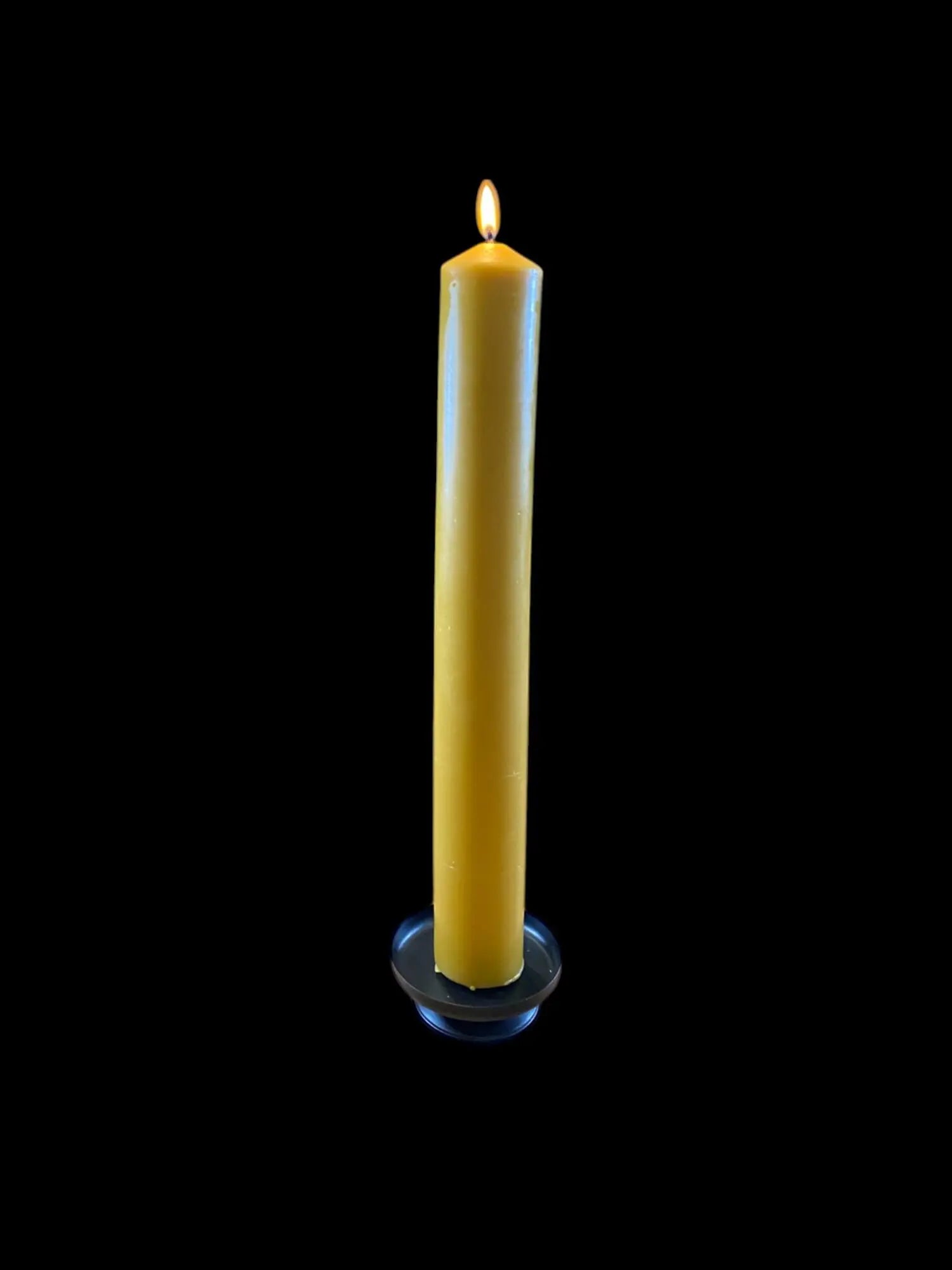 Tall lit beeswax church candle with natural honey color on black holder against black background