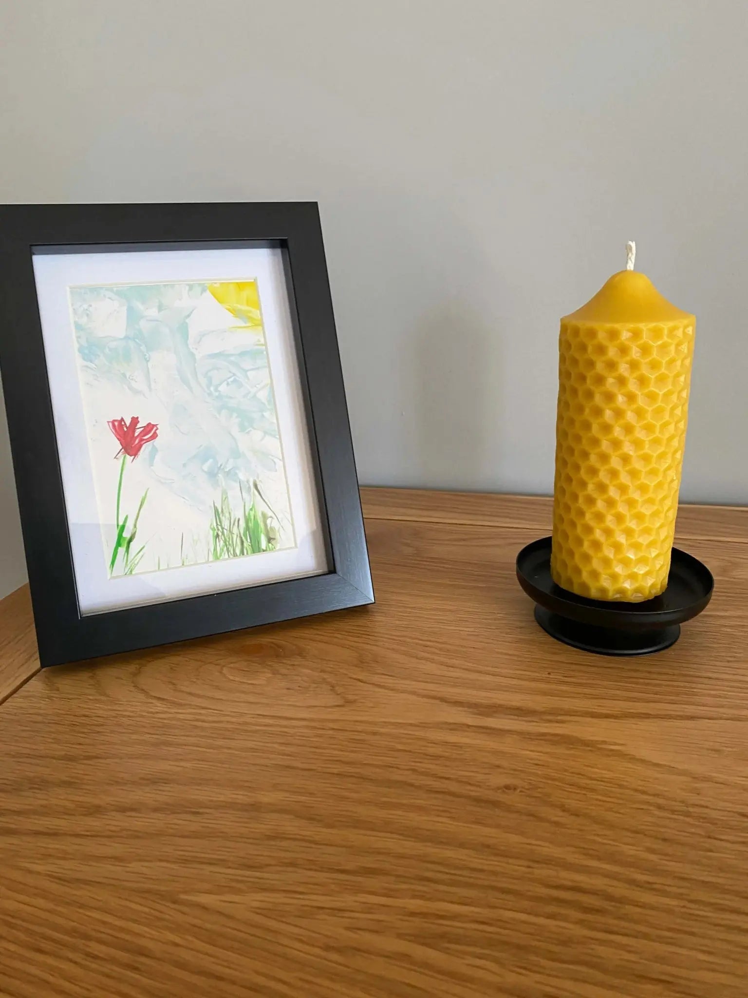 Handmade natural beeswax candle with honeycomb design on black holder beside framed floral watercolor art