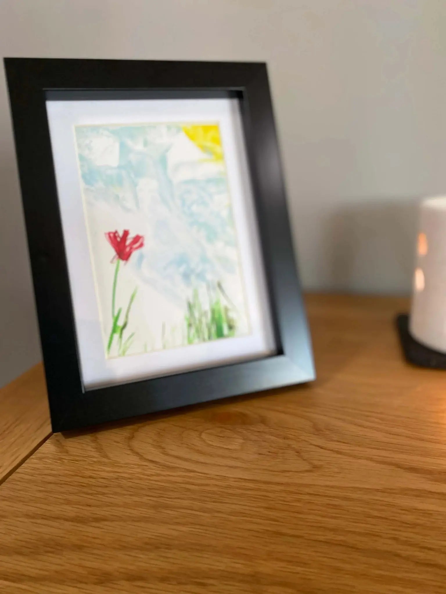 Framed artwork of a red flower with green stem and leaves against a light blue and yellow background on a wooden table