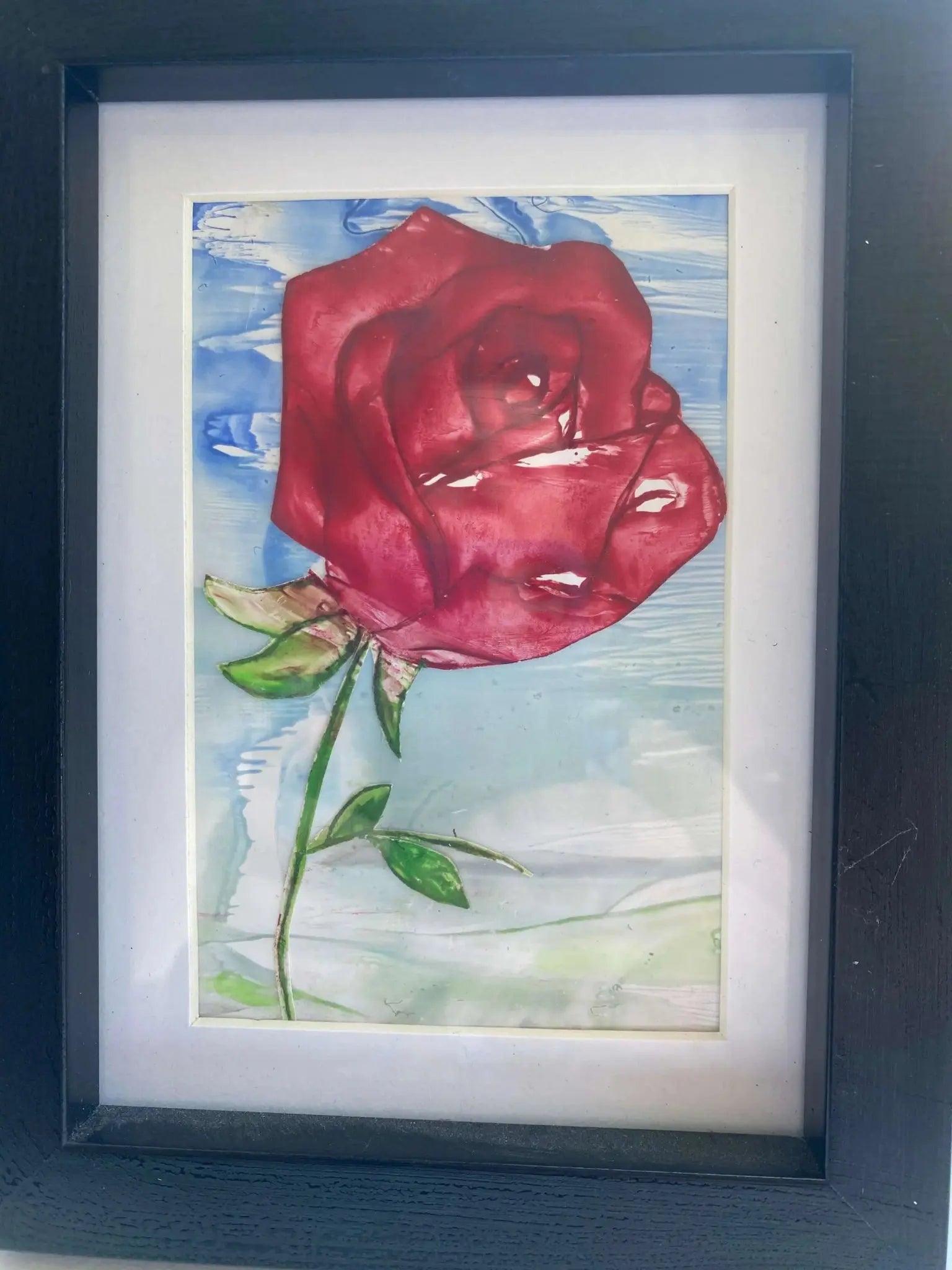 Framed encaustic art of a single red rose with green leaves against a blue and white background