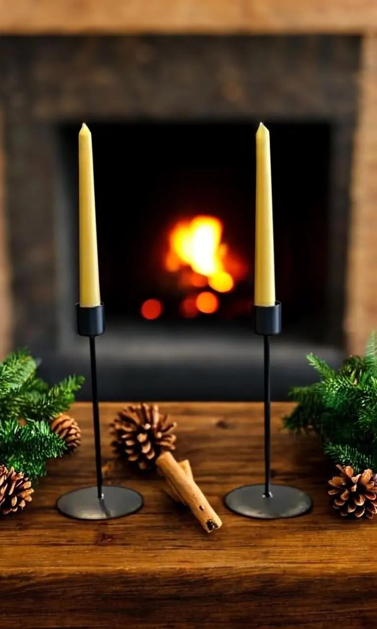 Pair of handmade taper beeswax candles in black holders with pine cones and greenery on wooden table in front of fireplace
