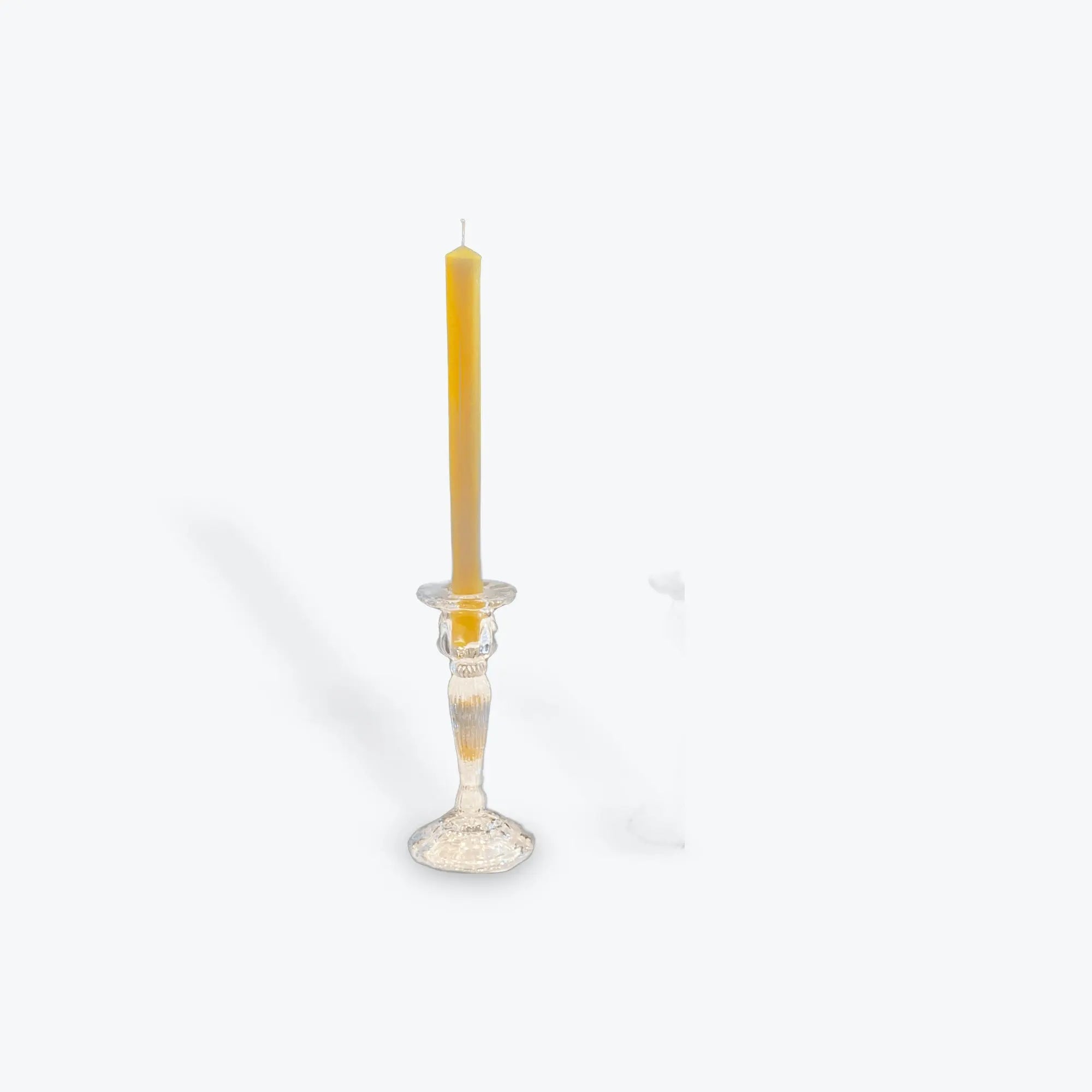 Single yellow beeswax taper candle in clear glass candlestick holder on white background
