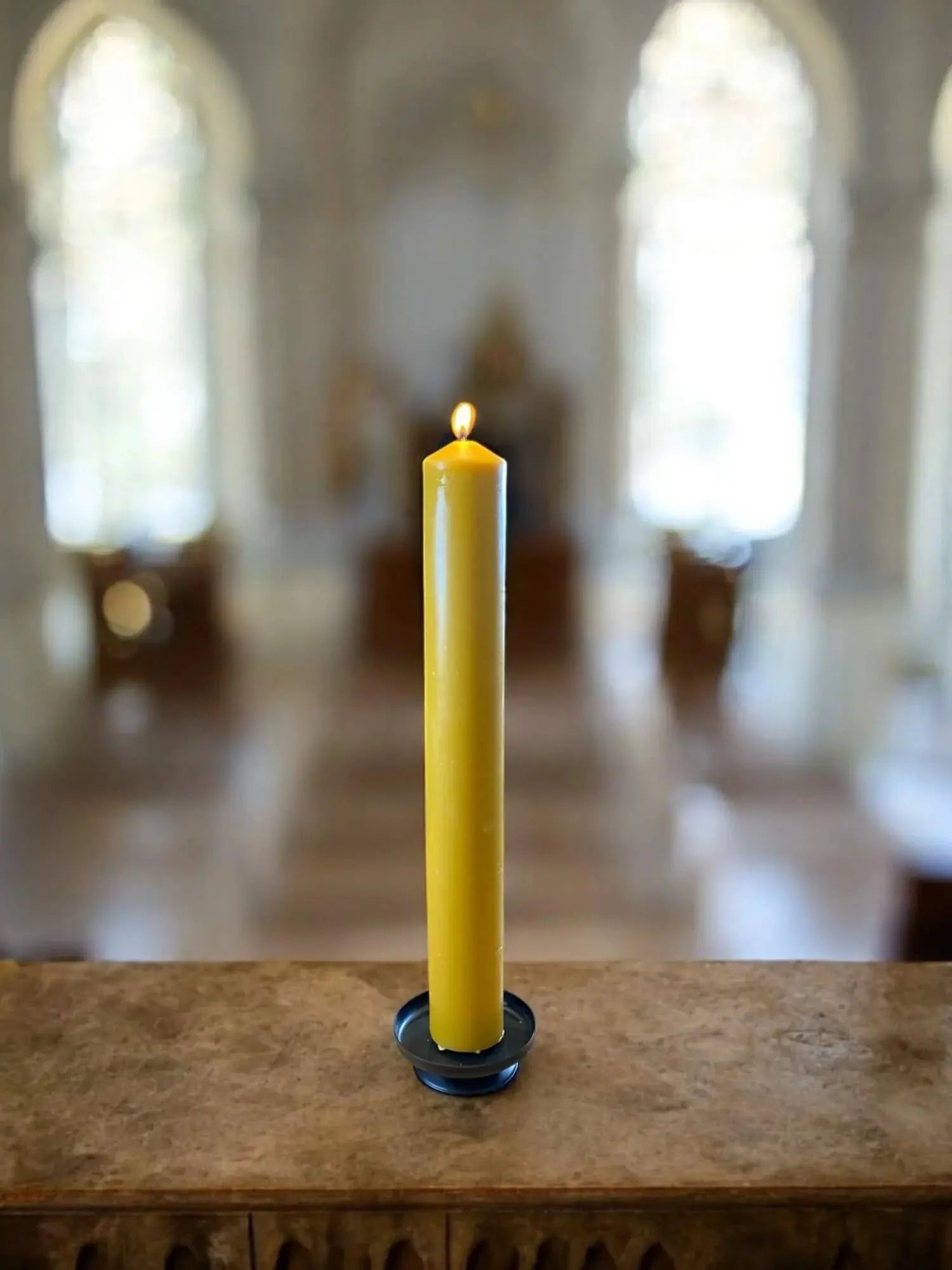 Tall lit yellow beeswax candle on a holder inside a softly lit church interior
